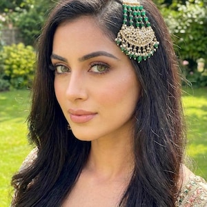 May include: A gold-toned hair accessory featuring a crescent-shaped pendant adorned with small white pearls and green beads. The piece includes five vertical strands of green beads and pearls that attach to the hair, creating a decorative effect.