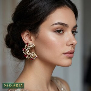 May include: A woman wearing a gold-coloured earring with red and clear gemstones. The earring has a crescent shape with an ornate design. The brand name "NOZARIA" is visible in the bottom left corner.