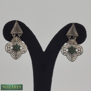 May include: Silver-toned earrings with a triangular top and a floral, openwork design. The earrings feature small, round, green gemstones at the center of the floral shape. The earrings are displayed on a black stand.