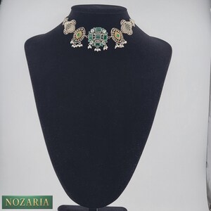 May include: Silver-toned choker necklace with green and yellow gemstone accents. The necklace features a central square design with smaller, circular side pieces. Small white beads hang from the bottom of the design. The necklace is displayed on a black mannequin.