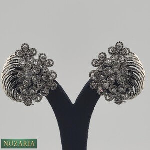 May include: A pair of silver-toned earrings with a floral design. Each earring features a cluster of small, round stones and a fan-shaped base with linear details. The earrings are displayed on a black stand against a neutral background.