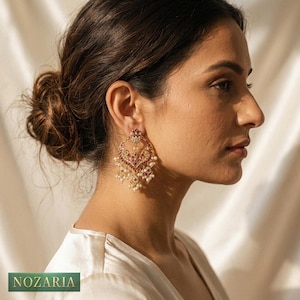 May include: Gold-toned earrings with a heart-shaped design, adorned with red gemstones and small white pearls. The earrings are hanging from the ear of a person. The brand name "NOZARIA" is visible at the bottom of the image.