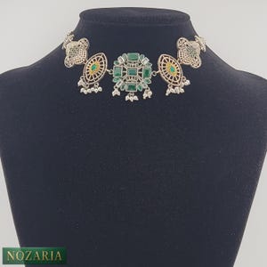May include: Silver-toned choker necklace with green gemstone accents. The necklace features a central cluster of square-cut green stones, flanked by oval and circular filigree designs. Small, dangling silver beads add detail. The necklace is displayed on a black mannequin.