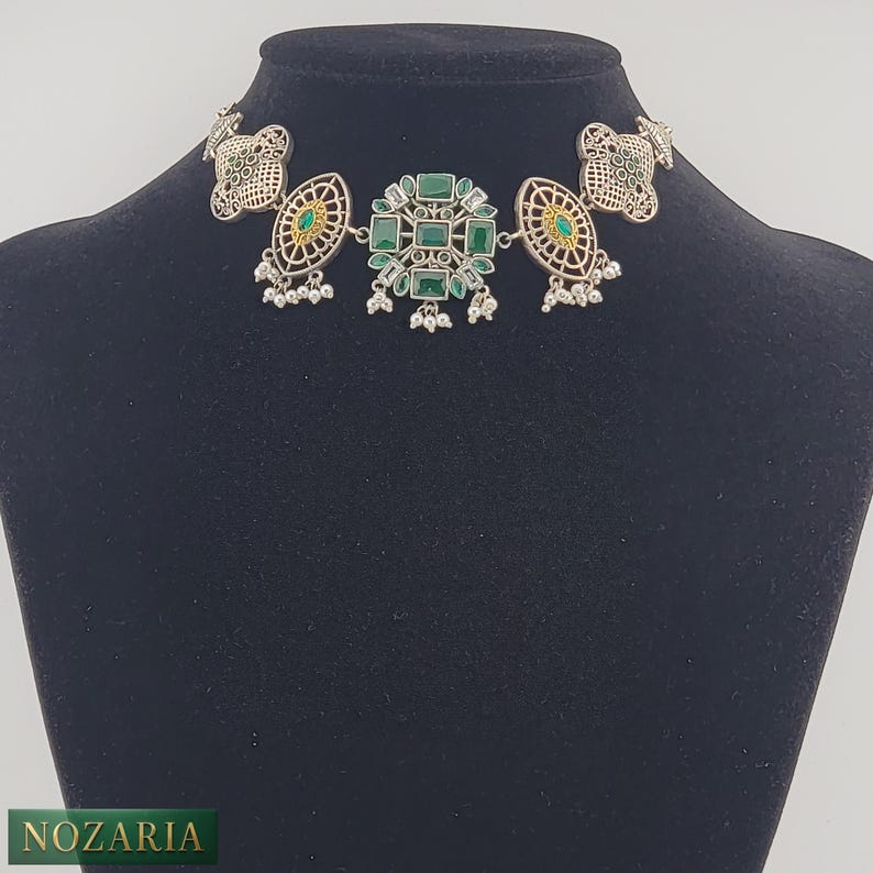 May include: Silver-toned choker necklace with green gemstone accents. The necklace features a central cluster of square-cut green stones, flanked by oval and circular filigree designs. Small, dangling silver beads add detail. The necklace is displayed on a black mannequin.