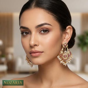 May include: Gold-coloured chandelier earrings featuring red gemstones and dangling pearls. The earrings have an ornate design with intricate detailing. The earrings are worn by a person with dark hair.