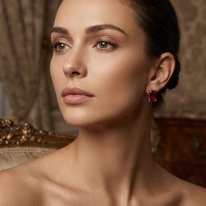 May include: A portrait of a woman wearing red rectangular gemstone earrings. The earrings are set in a silver or white metal. The image has a soft focus and the background is a blurred interior with ornate details. The brand name "NOZARIA" is at the bottom.