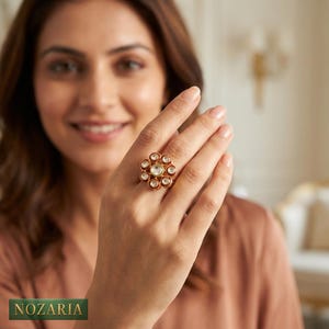 May include: A gold ring with a flower-like design, showcasing a central clear stone encircled by smaller, clear stones. The ring is worn on a hand, with the brand name "NOZARIA" displayed at the bottom of the image.