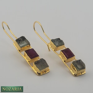 May include: Gold-tone earrings with three rectangular gemstones. The earrings feature a gold wire hook and a gold frame around each gemstone. The gemstones are a mix of gray and red hues. The brand name "NOZARIA" is at the bottom.