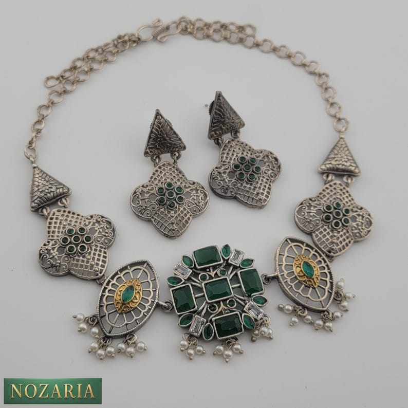 May include: A silver-toned necklace and earring set featuring intricate geometric designs and emerald-green gemstones. The necklace has a central cluster of square and rectangular stones, with smaller stones and pearl accents. The earrings mirror the necklace's design.