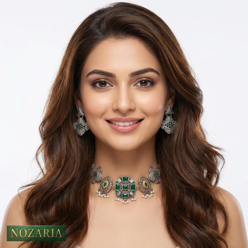 May include: A silver-toned necklace and earring set featuring green gemstone accents and intricate detailing. The necklace is a choker style with a central square design, complemented by matching earrings. The brand name "NOZARIA" is visible at the bottom.