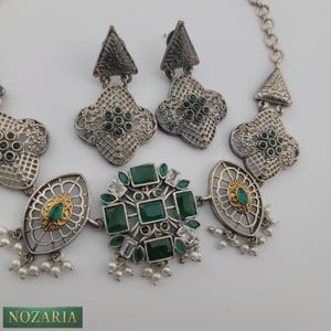 May include: Silver-toned jewelry set featuring a necklace and earrings. The necklace has a central cluster of green and clear stones, with oval and floral accents. The earrings have a matching floral design with green stones. The set includes small white pearl drops.