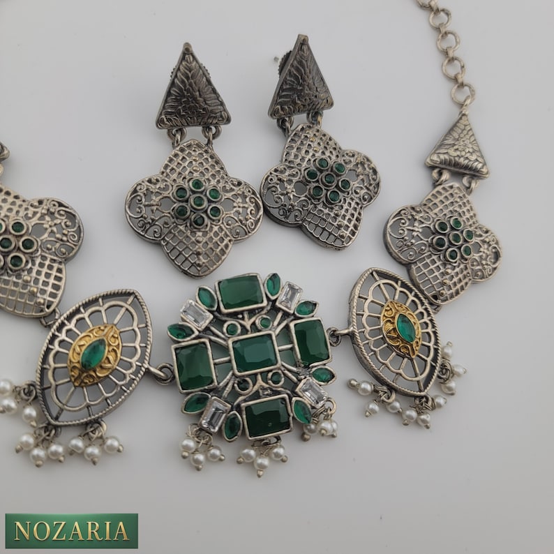 May include: Silver-toned jewelry set featuring a necklace and earrings. The necklace has a central cluster of green and clear stones, with oval and floral accents. The earrings have a matching floral design with green stones. The set includes small white pearl drops.
