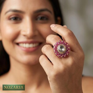 Peut inclure: Une bague ornée de grande taille avec une pierre centrale transparente entourée de petites pierres rouges vibrantes. La bague a un anneau et un sertissage de couleur or. La bague est sur le doigt d'une personne. La marque "NOZARIA" est en bas de l'image.