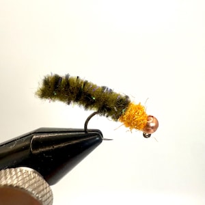 May include: A close-up of a handmade fishing fly. The fly features a dark green and black body with a copper-colored bead head and a bright orange collar. The fly is attached to a black hook.