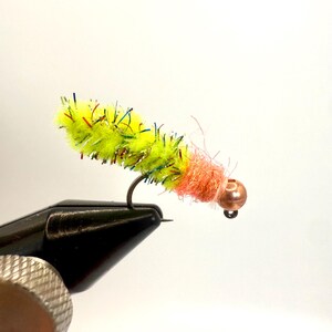 Może przedstawiać: Ręcznie wykonana przynęta muchowa z żółtym korpusem i pomarańczową główką. Korpus pokryty jest kolorowymi, opalizującymi włóknami. Haczyk jest czarny, a główka ma miedzianą koralik.