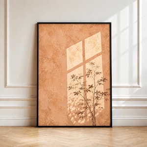 May include: A rectangular wall art print in a black frame featuring a textured terracotta-colored background. The image displays a shadow of a window pane casting a cross shape over the surface, overlaid with the silhouette of a plant.