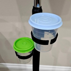 Puede incluir: Dos recipientes de plástico transparente con tapas, uno azul claro y otro verde, están sujetos a un poste negro con correas negras. Los recipientes parecen ser de la marca Tupperware. El fondo es una pared beige neutra.