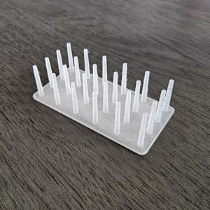 May include: A small, rectangular, white plastic object with numerous vertical, cylindrical pegs. The pegs are evenly spaced and protrude upwards from the base. The object is set against a dark, wood-grain surface.