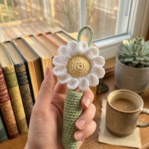 May include: A handmade crochet bookmark featuring a white daisy with a yellow center and a green stem. The bookmark is held in a hand, with a background of books, a succulent plant, and a coffee mug.