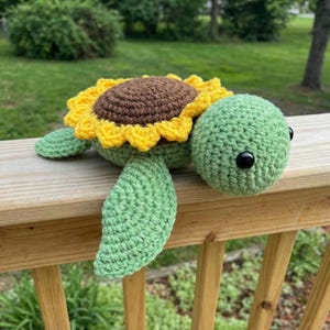May include: A handmade crochet turtle with a sunflower design. The turtle's head and body are green, with a brown center and yellow petals forming the sunflower. The turtle has black eyes and green flippers.