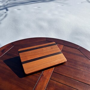 May include: A square wooden cutting board with a light brown base and two dark brown stripes. The board is displayed on a dark brown wooden table, with a snowy background.