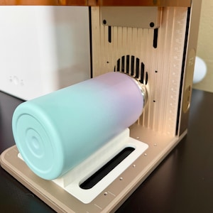 May include: A tumbler with a gradient design in shades of blue and purple is placed inside a laser engraving machine. The machine has a gold-colored frame and a clear orange top. The tumbler is resting on a white platform.