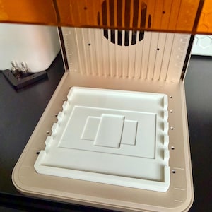 May include: A resin 3D printer build plate featuring a metallic gold base with vertical alignment markings and a white rectangular platform with raised side edges and a central recessed rectangular design, topped by an amber-colored cover.