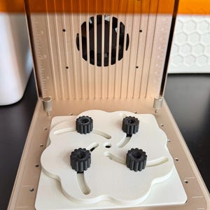 May include: An open 3D printer with a beige frame and orange top. The printer's interior features a white platform with four black, gear-shaped knobs. A fan is visible in the upper section.