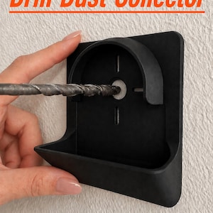 May include: Black drill dust collector with the text "Drill Dust Collector" in orange. The tool is held against a white textured wall, with a drill bit inserted into the collector. The collector has a curved top and a tray-like bottom.