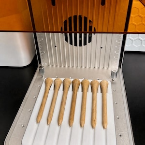 May include: A close-up shot of a 3D printer with a series of wooden golf tees. The tees are light brown, with a rounded top and a pointed end. The printer has a silver metal base and a clear orange cover.
