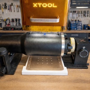 May include: A black and gold XTOOL laser engraving machine is displayed on a wooden surface. The machine has a cylindrical body and a rectangular base with a grid of holes. The word "XTOOL" is illuminated in gold.