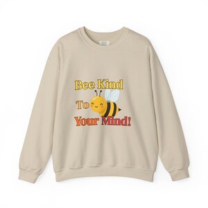 May include: A cream-coloured sweatshirt featuring the phrase "Bee Kind To Your Mind!" in yellow and red. The design includes a cartoon bee with a smiling face and black and yellow stripes. The sweatshirt has long sleeves and a crew neck.