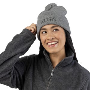Peut inclure: Un bonnet en tricot gris avec un pompon sur le dessus et le mot "LOVE" brodé en script noir. Le bonnet est porté par une personne portant une veste polaire grise.