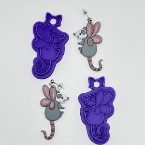 May include: Two purple plastic cookie cutters shaped like a fairy opossum with a bow at the top, alongside two matching earrings featuring a gray opossum with pink glittery wings and a long tail, each with a silver-toned metal earring post.