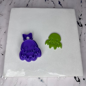 May include: Two octopus-shaped cookie cutters. One is purple with a bow, the other is green. The cutters are made of plastic and are designed for making cookies or other treats. The cutters are on a white surface.