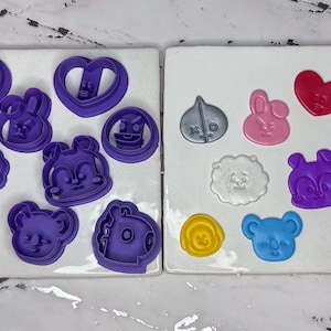 May include: A collection of purple and colored cookie cutters in various shapes, including animals, hearts, and flowers. The cutters are arranged on two white rectangular tiles. The colors include purple, pink, silver, yellow, brown, and blue.