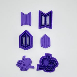 Puede incluir: Un juego de seis cortadores de galletas de plástico morado dispuestos en tres pares. Las formas incluyen dos contornos de logotipo trapezoidales, dos contornos de logotipo hexagonales y dos contornos de logotipo de corazón entrelazados.
