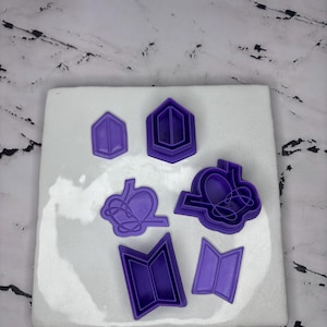 May include: A collection of purple plastic cookie cutters in various shapes, including a hexagon, a heart with a crown, and an open book. The cutters are arranged on a white surface with a marble-like background.