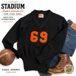 Puede incluir: Suéter negro estilo universitario con el número 69 en naranja. El suéter está hecho de 100% lana pura y tiene cuello en V. La imagen también incluye un balón de fútbol, jeans y botas de cuero marrón. El texto en el suéter dice "Stadium Shaker Sweater Co."