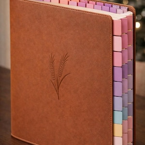 Może przedstawiać: Brązowy, skórzany notatnik z wytłoczonym wzorem kłosa pszenicy na okładce. Notatnik posiada kolorowe zakładki w odcieniach różu, fioletu i błękitu. Krawędzie są zaokrąglone, a notatnik wydaje się nowy.