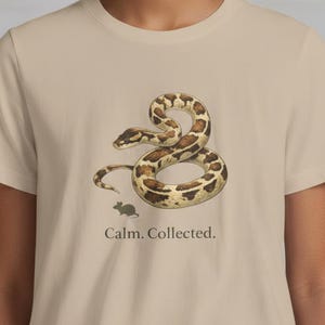 May include: Beige t-shirt with a coiled snake illustration in brown and tan, and a small mouse. The text "Calm. Collected." is printed below the graphic. The shirt has short sleeves and a crew neck.