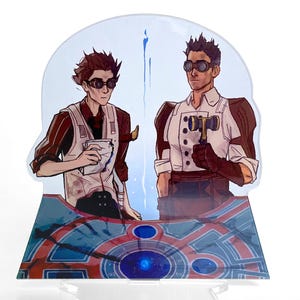 以下が含まれることがあります： スチームパンク風のイラストが描かれた、透明なアクリルスタンド。2人の人物が描かれており、一人はカップを、もう一人はハンマーを持っています。ベースは青と赤の幾何学模様で、中央に青い光があります。