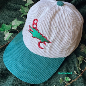 May include: White corduroy baseball cap with a teal brim and button. The cap features a green alligator design with red accents. A stylish accessory.