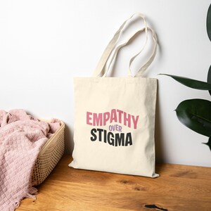 May include: Cream-colored tote bag with the words "EMPATHY OVER STIGMA" in pink and black. The bag has long handles and is made of a sturdy fabric. A pink blanket is in a basket to the left of the bag.