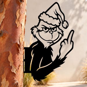 May include: A black metal silhouette wall art piece depicting a grumpy character wearing a Santa hat, with a furrowed brow and a smirk, holding up a middle finger. The cutout design features sharp edges and is mounted against a textured tree trunk.
