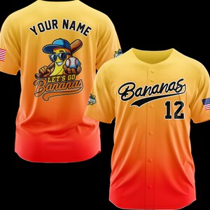 Puede incluir: Dos camisetas de b&eacute;isbol. Una es amarilla y naranja con "Your Name" en la espalda y un personaje de pl&aacute;tano con gafas de sol, un bate de b&eacute;isbol y una pelota de b&eacute;isbol. La otra camiseta es amarilla y naranja con "Bananas" y el n&uacute;mero "12".