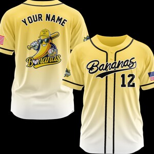K&ouml;nnte beinhalten: Zwei gelbe Baseball-Trikots. Eines hat "YOUR NAME" auf dem R&uuml;cken und ein Bananen-Maskottchen mit Sonnenbrille und Baseballschl&auml;ger. Das andere Trikot hat "Bananas" und die Nummer "12" auf der Vorderseite. Beide haben schwarze Akzente und amerikanische Flaggen.