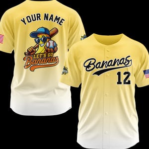 Puede incluir: Dos camisetas de b&eacute;isbol amarillas. Una presenta un personaje de pl&aacute;tano con gafas de sol, un bate de b&eacute;isbol y el texto "Let's Go Bananas". La otra camiseta tiene "Bananas" y el n&uacute;mero "12" en la parte delantera. Ambas tienen una bandera estadounidense.