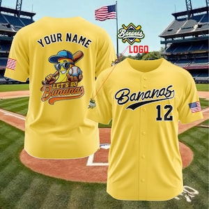 Puede incluir: Camiseta de b&eacute;isbol amarilla con la palabra "Bananas" en escritura negra y el n&uacute;mero 12. Un parche de la bandera estadounidense est&aacute; en la manga. La parte posterior de la camiseta tiene "Your Name" encima de un personaje de pl&aacute;tano de dibujos animados con un bate y una pelota de b&eacute;isbol.