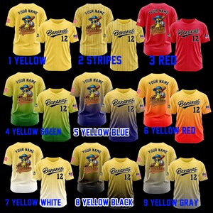 Puede incluir: Una colecci&oacute;n de camisetas de b&eacute;isbol en varios colores, incluyendo amarillo, rojo y negro. Cada camiseta presenta el texto "Bananas 12" y un personaje de pl&aacute;tano de dibujos animados con un bate de b&eacute;isbol. Las camisetas se muestran con el texto "YOUR NAME" en la parte superior.